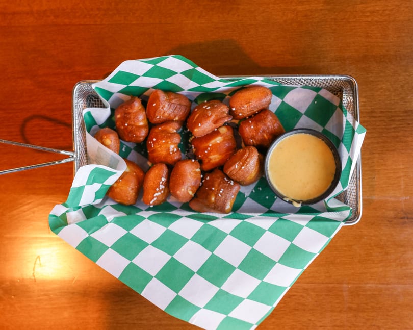 Pretzel bites with salt and a side of cheese dip in a metal basket with green and white checkered paper.