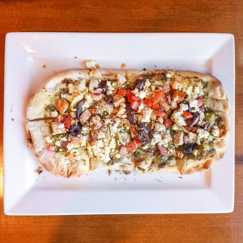 Overhead view of a rectangular flatbread pizza with pesto, feta, chicken, olives, and tomatoes.