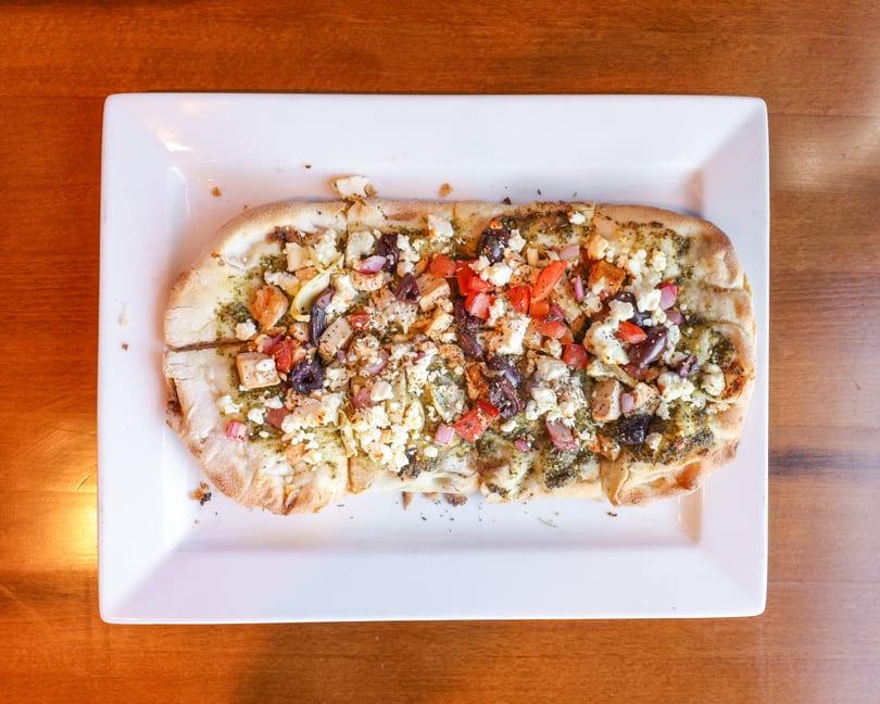 Overhead view of a rectangular flatbread pizza with pesto, feta, chicken, olives, and tomatoes.
