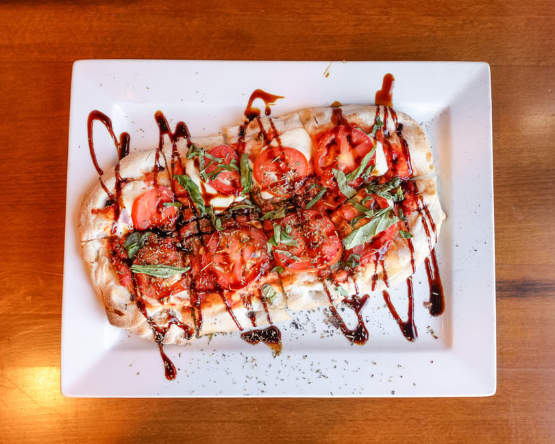 Caprese flatbread with sliced tomatoes, mozzarella, basil, and balsamic glaze on a white rectangular plate.