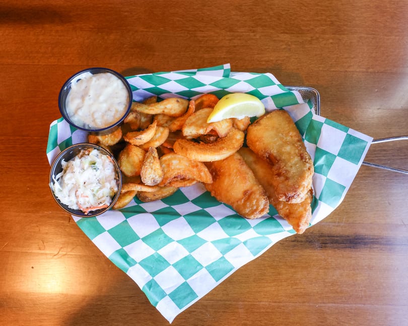 Fish and chips with coleslaw, tartar sauce, and a lemon wedge in a basket on a wooden table.