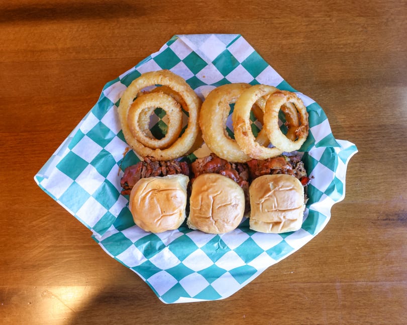 Three sliders with meat and sauce, topped with crispy onion rings, served in a checkered paper basket.