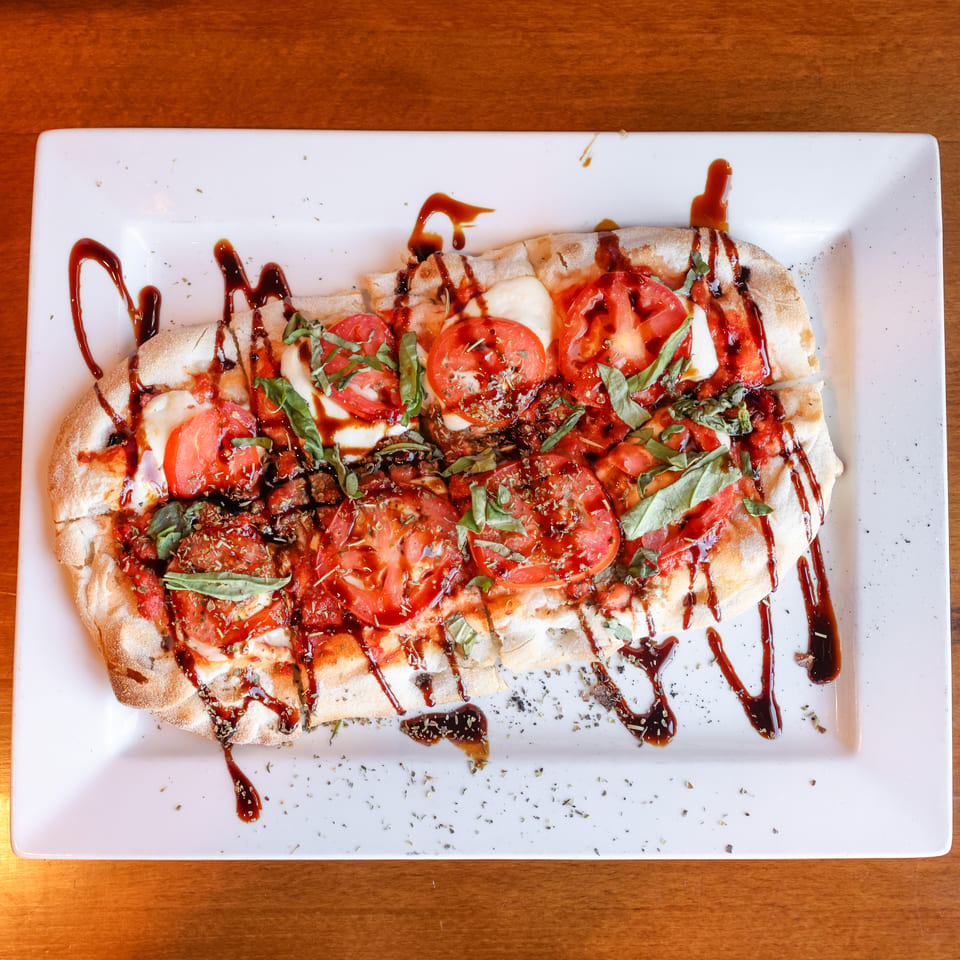 Caprese flatbread with sliced tomatoes, mozzarella, basil, and balsamic glaze on a white rectangular plate.