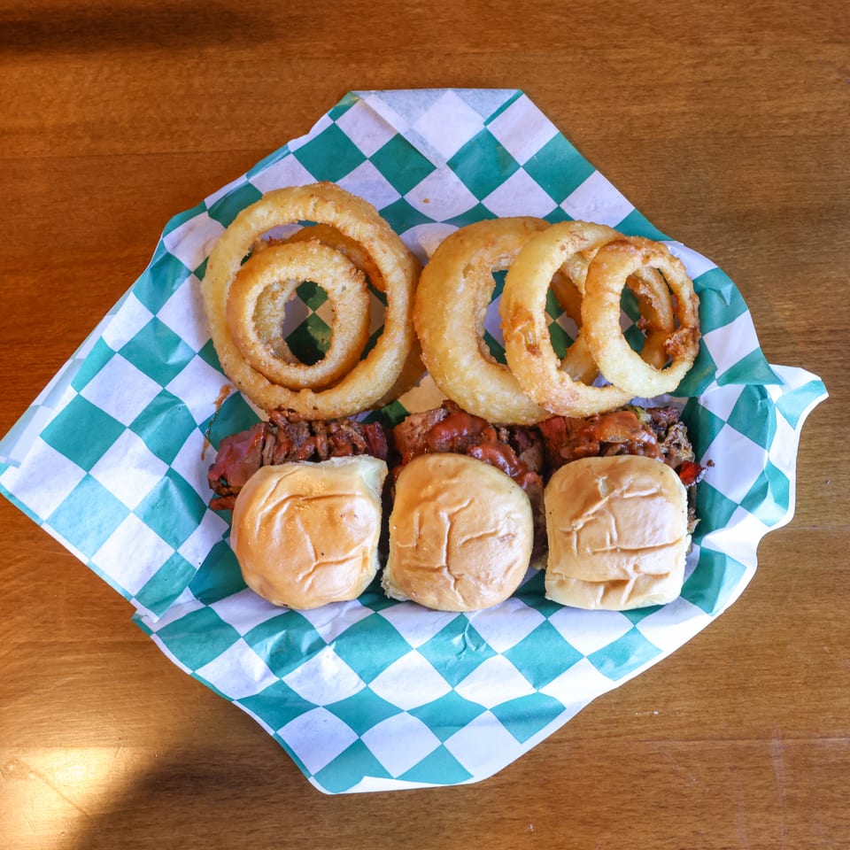 Three sliders with meat and sauce, topped with crispy onion rings, served in a checkered paper.