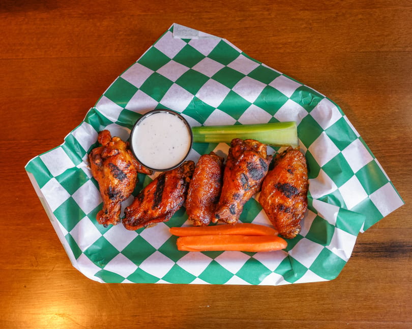 Grilled chicken wings with ranch dressing, celery, and carrots in a green checkered paper-lined basket.