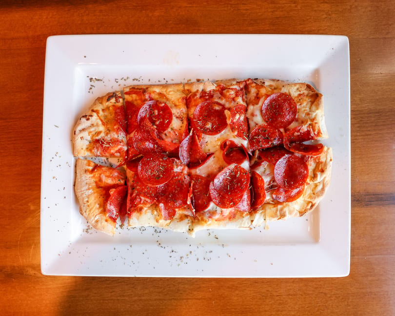 Pepperoni flatbread pizza, cut into squares, on a white rectangular plate on a wooden table.