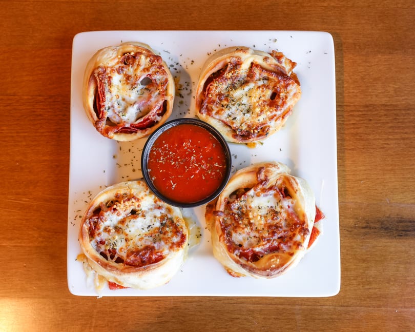 Four baked pepperoni and cheese pizza rolls with a side of marinara sauce on a white plate.