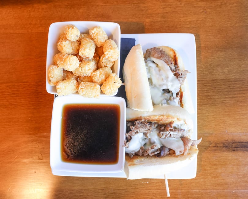 Overhead view of a Philly cheesesteak sandwich, tater tots, and au jus on a wooden table.